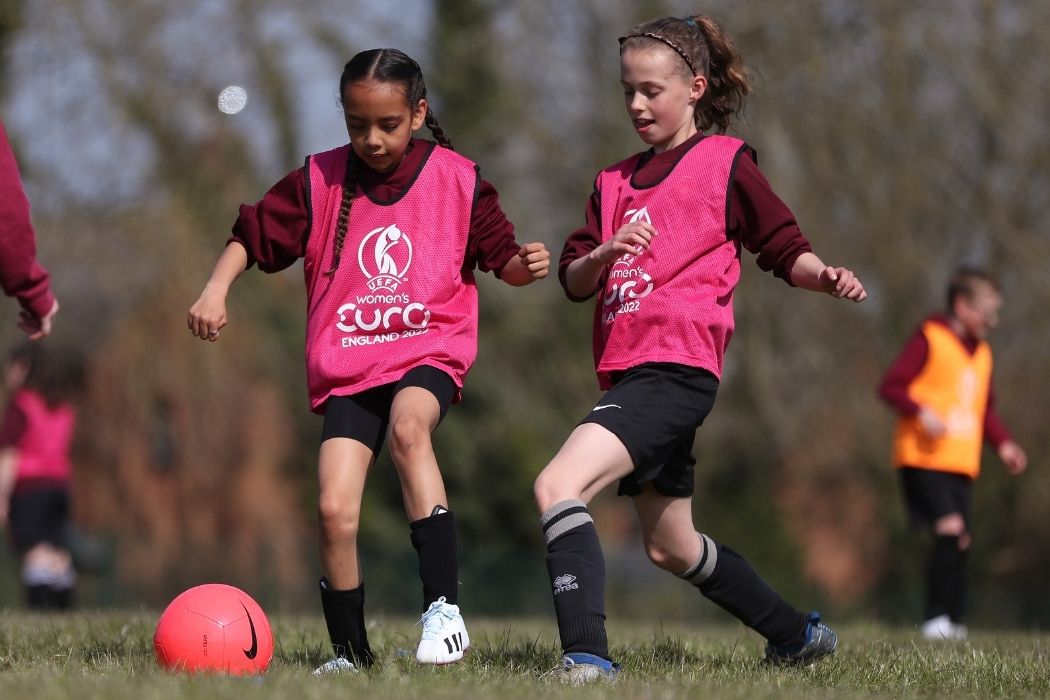 Two girls playing football. They are wearing pink vests which have 'UEFA Women's EURO England 2022' written on them.