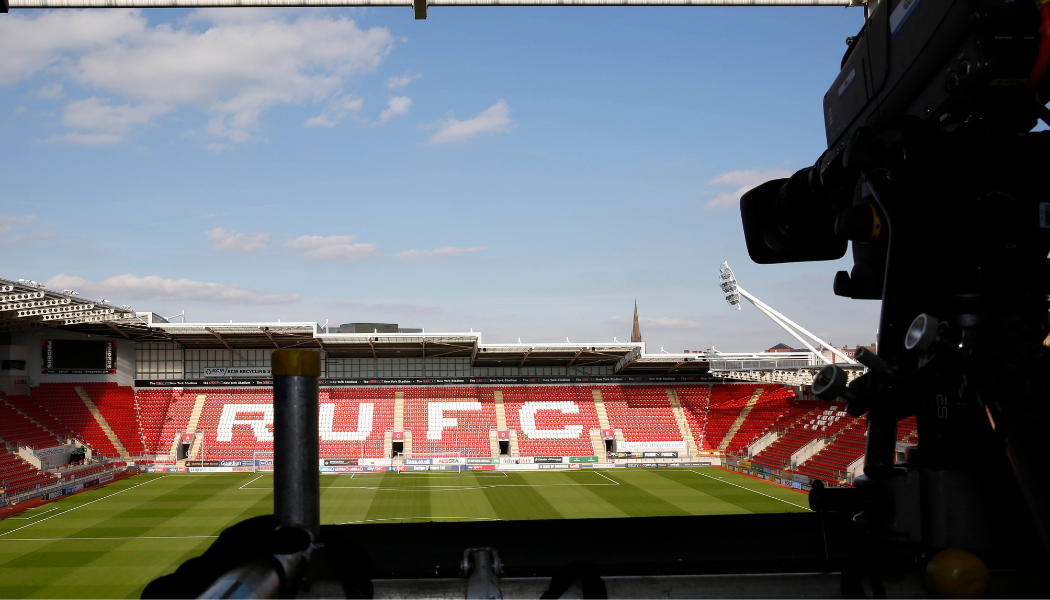 A camera pointing towards a football stadium stand that has RUFC displayed on it.